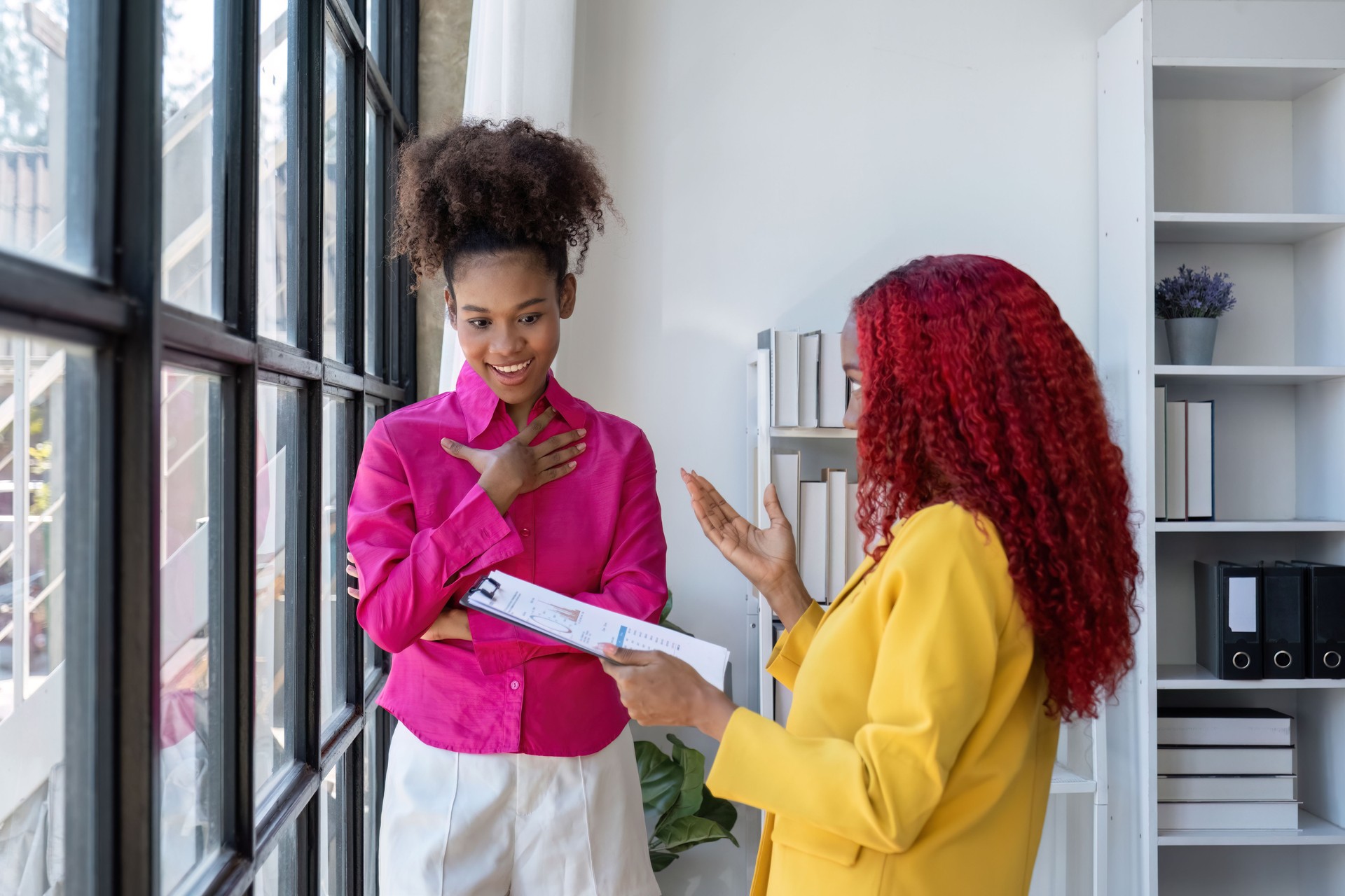 Two African American businesswomen using laptop and clipboards stand talking, giving advice on work and planning accounting and finance work together in the office.